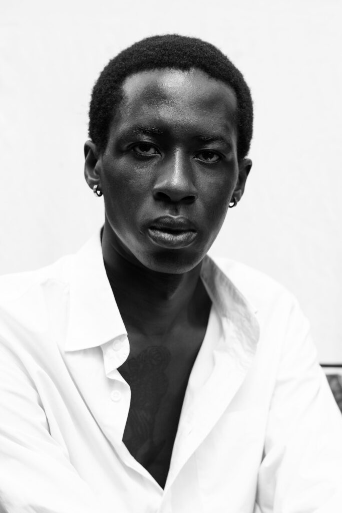 Black and white studio portrait of a man in an open white shirt against a plain wall
