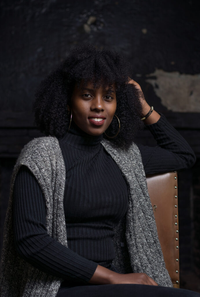 Indoor portrait of a woman in a black turtleneck and grey knit waistcoat seated on a leather chair