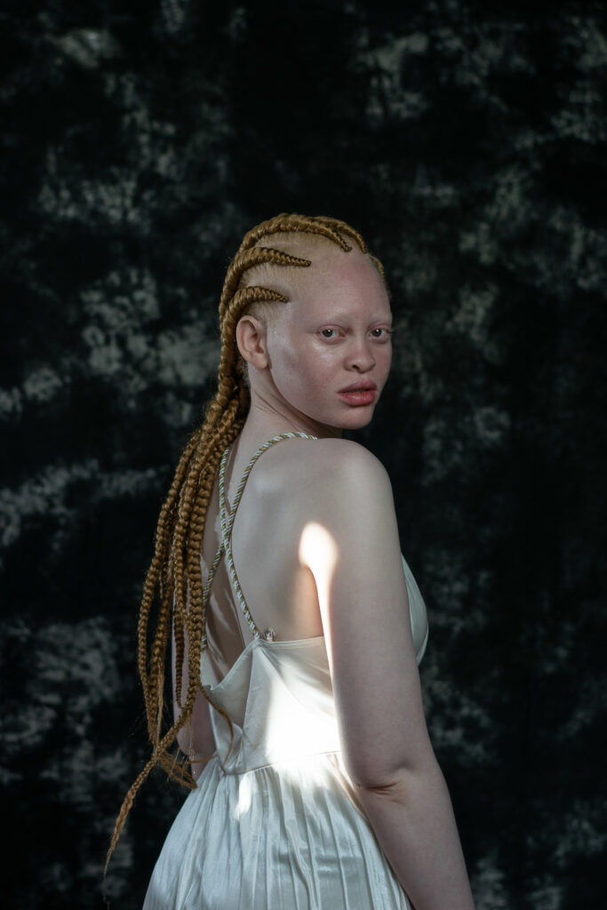 Fine art portrait of a woman with long braids in a pale slip dress, photographed from behind against a dark backdrop